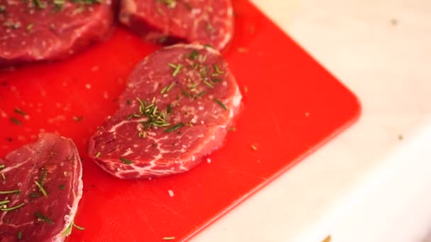 Five raw meat pieces of steak on a red cutting board, sprinkled with rosemary and coarse salt. — Stock Video