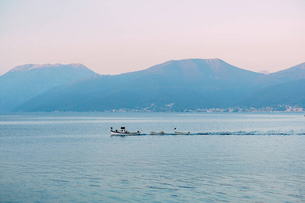 White motor boat tows small rowboats on the sea along the mountains at sunset