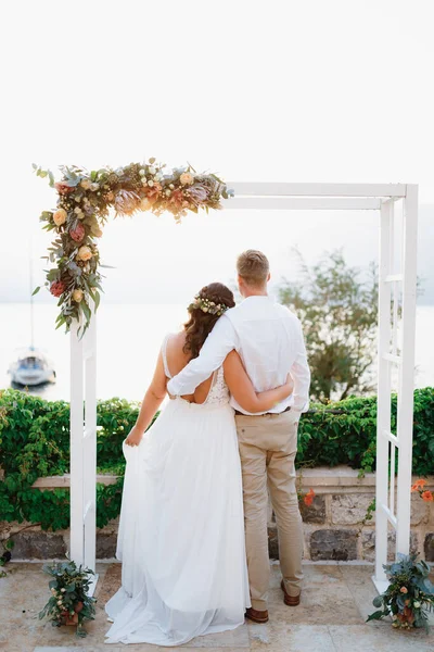 A noiva e o noivo se abraçam sob o arco do casamento e olham para o mar, vista traseira — Fotografia de Stock