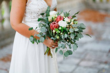 Nazik bir gelin elinde beyaz şakayıklardan, güllerden, proteinlerden ve okaliptüslerden oluşan sıradışı bir buket tutuyor. 