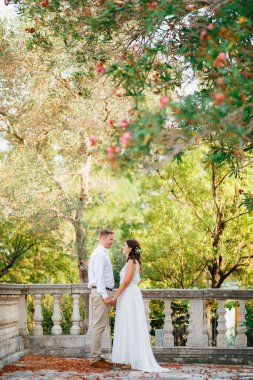 Gelin ve damat tırabzanın yanında duruyorlar çiçek açan bir ağacın dallarının altında el ele tutuşuyorlar ve birbirlerine bakıyorlar. 