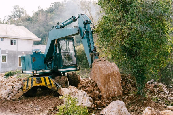 Blue excavator works at a construction site against the backdrop of a house and greenery