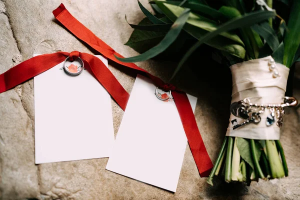 Cincin pernikahan yang diikat dengan pita merah terletak pada kartu kosong dengan hiasan di samping karangan bunga dengan latar belakang ubin batu — Stok Foto
