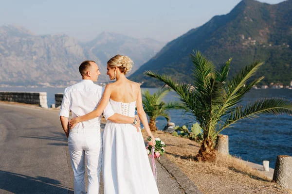 Braut und Bräutigam spazieren entlang der Seebrücke in der Nähe der Altstadt von Perast, umarmen sich, Rückansicht — Stockfoto