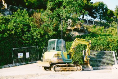 Parkta metal bir çitin arka planında çalışan kovalı sarı bir traktör.