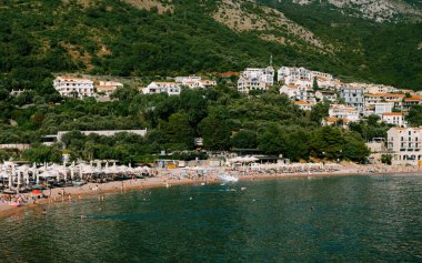 Karadağ 'ın Sveti Stefan adası yakınlarındaki antik kentin arka planında güneşli şezlonglar ve şemsiyelerle sahilin manzarası