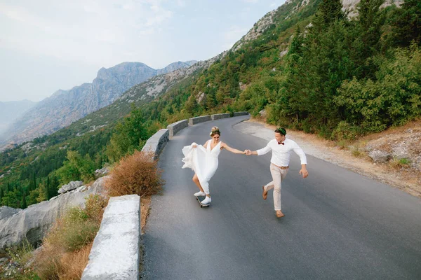 신부가 스케이트보드를 타고 산속 도로를 따라 달리는 신랑의 손을 잡고 있다 — 스톡 사진