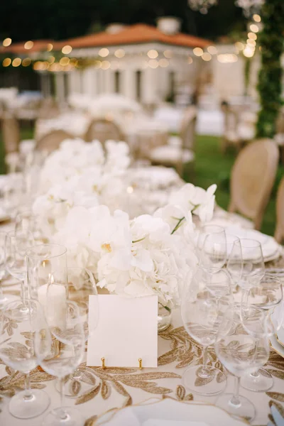 La tarjeta vacía se apoya sobre una mesa cubierta con un mantel blanco sobre el fondo de un ramo de flores y vasos vacíos — Foto de Stock