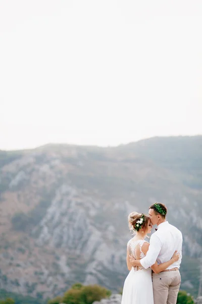 Lo sposo e la sposa in ghirlande stanno sulla cima del monte abbracciando, lo sposo bacia la sposa sulla fronte — Foto Stock