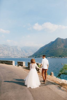 Gelin ve damat, Perast yakınlarındaki Kotor Körfezi kıyısında yol boyunca el ele yürür.