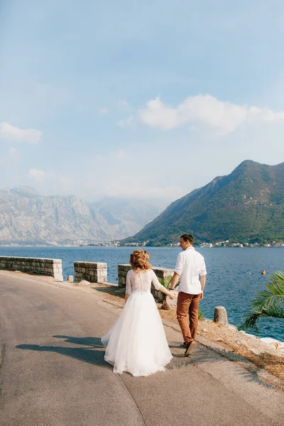 Panna młoda i pan młody idą ręka w rękę wzdłuż drogi wzdłuż wybrzeża w Zatoce Kotor w pobliżu Perast, widok z tyłu — Zdjęcie stockowe