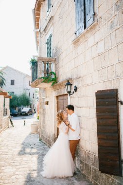 Gelin ve damat, eski Perast kasabasında balkonları ve ahşap panjurları olan güzel beyaz bir binanın yanında genç bir ağacın yanında sarılıyorlar. 