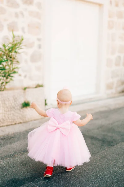 Una niña encantadora en un vestido rosa con un arco, zapatillas de deporte y una diadema corre a lo largo de la carretera en el casco antiguo, vista trasera —  Fotos de Stock