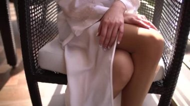 Çıplak ayaklı kadın, ipek bornozlu, balkonda sandalyede, elleri dizlerinin üzerinde.