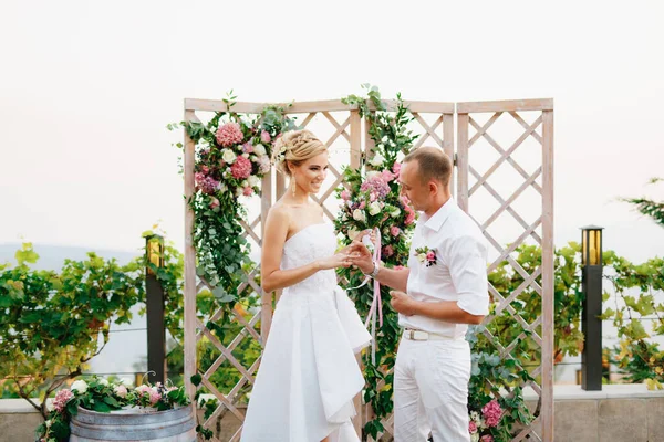 Braut und Bräutigam stehen in der Nähe des Hochzeitsbogens, der Bräutigam legt den Ring während der Trauung an den Finger der Braut — Stockfoto