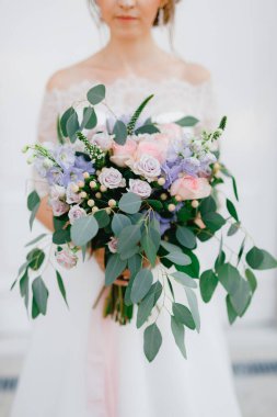 Yumuşak bir gelin elinde güller, Veronica, Delphinium ve okaliptüs dallarından oluşan bir buket tutuyor. 