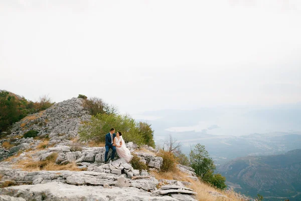 Braut und Bräutigam sitzen umarmt auf einem Felsen auf dem Berg Lovcen, hinter ihnen liegt die Bucht von Kotor — Stockfoto