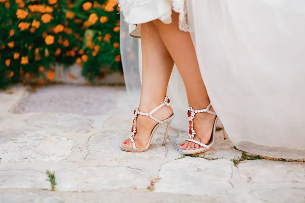Brudben i snygga sandaler med kristaller med höga klackar kika ut under brudklänningen, närbild — Stockfoto