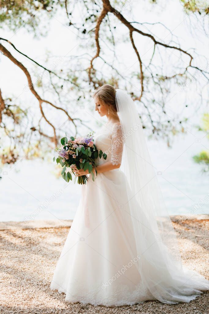  A sophisticated bride stands with a wedding bouquet in her hands under the branches of a tree by the sea 