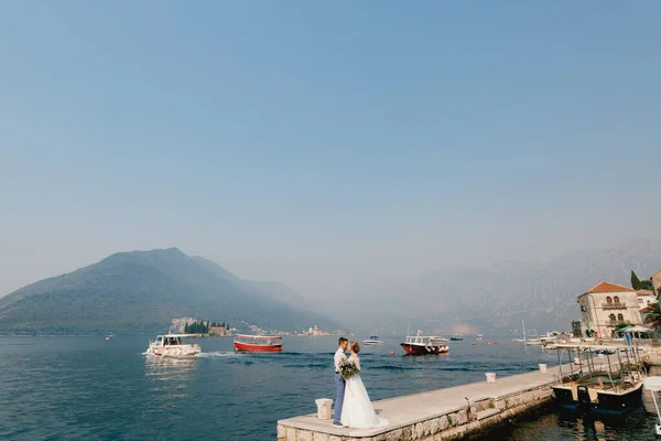 A noiva eo noivo estão abraçando no cais perto da cidade velha de Perast, barcos estão flutuando nas proximidades — Fotografia de Stock