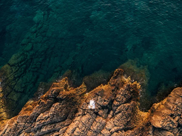 La novia y el novio se encuentran abrazados en una orilla rocosa, frente a ellos hay un mar azul profundo, vista superior — Foto de Stock