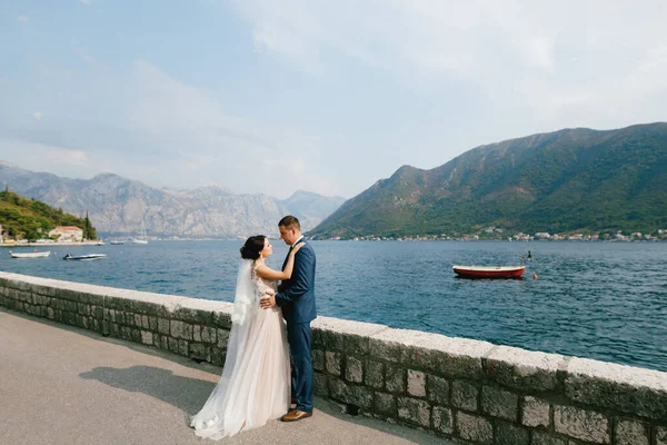 The bride and groom hugging on the pier near old town of Perast, behind them is a small red boat — Stock Photo, Image