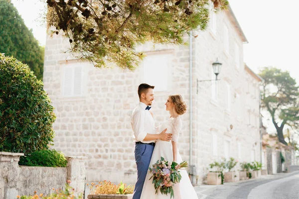 Bruden och brudgummen står kramas under tallkvistar nära en vacker tegelhus i den gamla staden Perast — Stockfoto