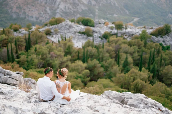 Braut und Bräutigam sitzen umarmt auf einem Felsen auf dem Lovcen und schauen einander an, Rückansicht — Stockfoto