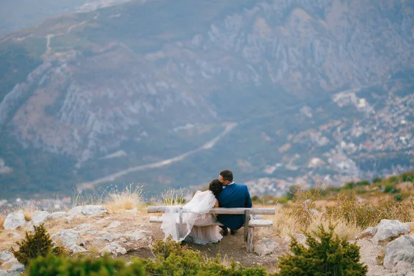De bruid en bruidegom zitten teder op een houten bankje in de bergen — Stockfoto