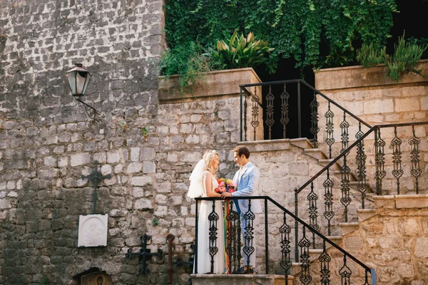 Nevěsta a ženich stojí na krásném schodišti s tepaným železným zábradlím ve starém městě Perast a dívají se na sebe — Stock fotografie