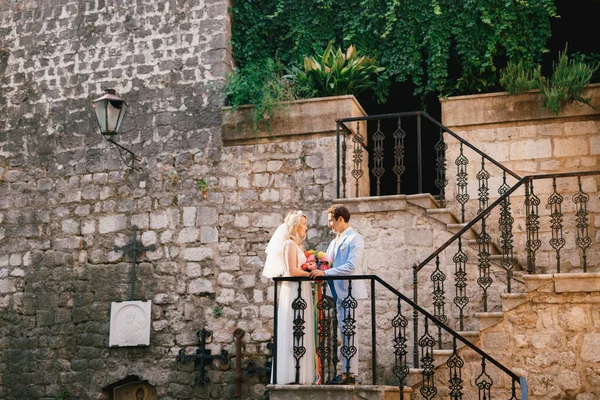 La novia y el novio se paran en una hermosa escalera con una barandilla de hierro forjado en el casco antiguo de Perast y se miran entre sí —  Fotos de Stock