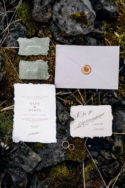 Wedding rings lie on moss-covered stones, surrounded by decorated invitations and envelope sealed with sealing wax. Nearby are two signs with inscriptions. Inscription: groom, bride — Stock Photo, Image