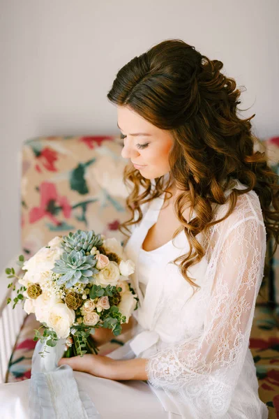 Μια νύφη με δαντελωτή ρόμπα κάθεται σε έναν καναπέ σε ένα δωμάτιο ξενοδοχείου και κρατά ένα γαμήλιο μπουκέτο στα χέρια της, κοντινό πλάνο — Φωτογραφία Αρχείου