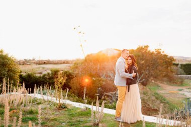 Takım elbiseli, gülümseyen adam pastel elbiseli ve siyah deri ceketli ciddi bir kıza sarılıyor.