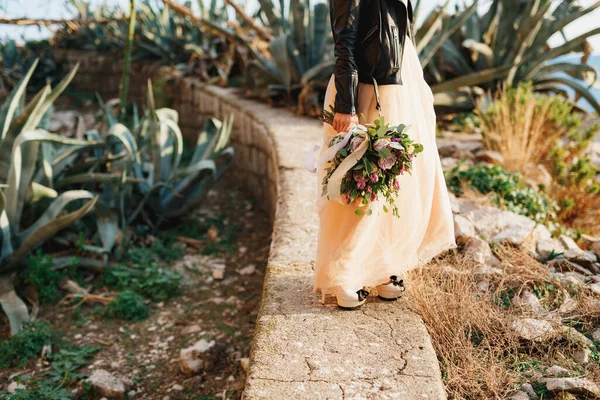 Pastel elbisesi ve deri ceketli gelin bir buket çiçekle bir yolda duruyor. — Stok fotoğraf