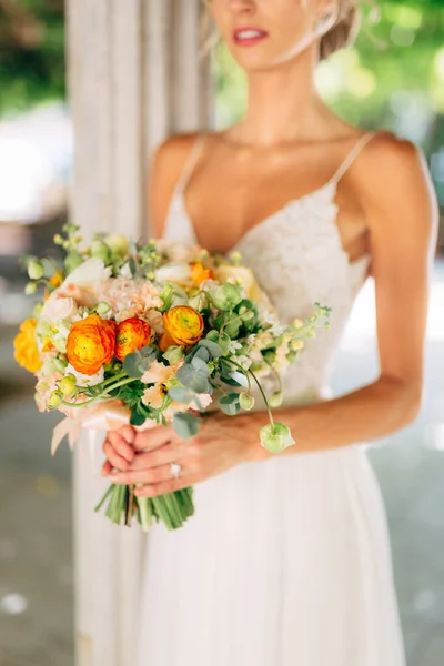 La novia se encuentra entre columnas blancas trenzadas con vides verdes y sostiene un ramo de bodas con botones de naranja, primer plano — Foto de Stock