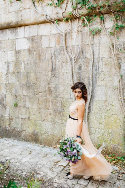 Vacker brud i en pastell klänning och högklackade skor promenader med en lyxig bukett blommor längs en stenmur — Stockfoto