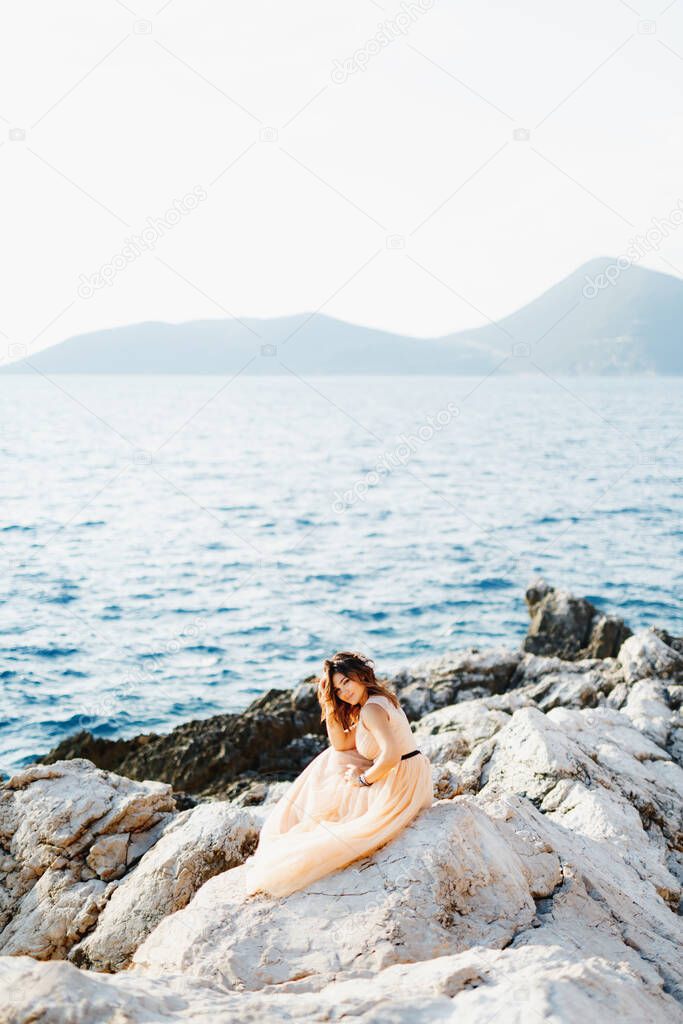 Smiling bride with beautiful makeup sits in a pastel wedding dress on a rock above the sea against a background of mountains