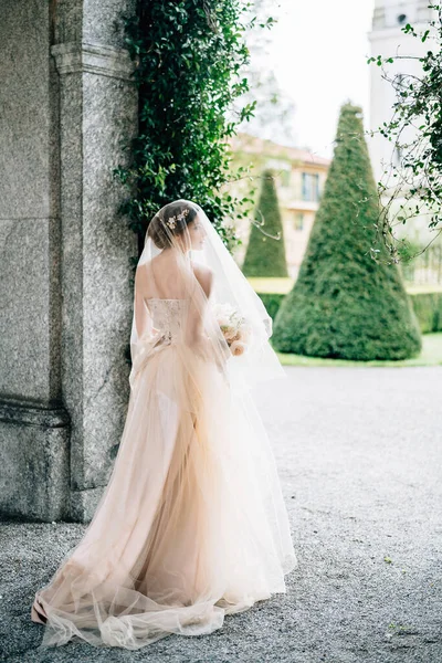 Novia en un vestido de novia con un velo y un ramo de flores se encuentra cerca del arco de un antiguo edificio frente a un jardín verde. Lago de Como —  Fotos de Stock