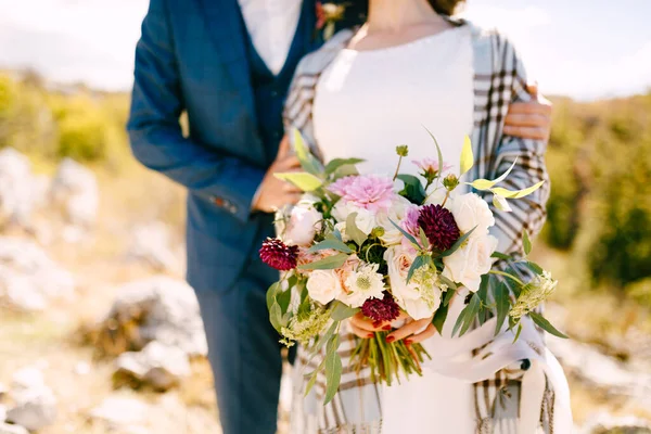 Groom étreint les épaules de la mariée dans un châle à carreaux avec un beau bouquet de fleurs dans ses mains — Photo