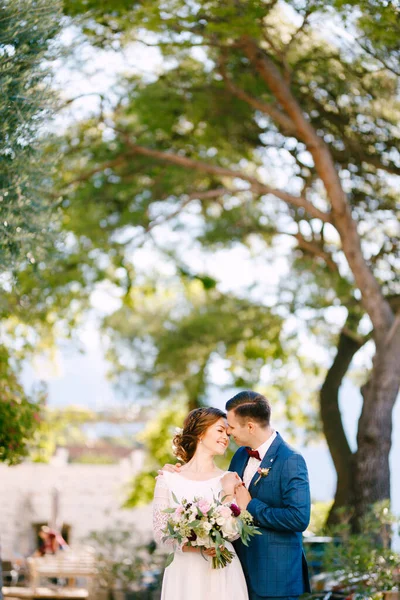 Ο γαμπρός αγκαλιάζει απαλά τη νύφη από τους ώμους με φόντο τα δέντρα σε μια ζεστή ηλιόλουστη μέρα. Η νύφη κρατά ένα όμορφο μπουκέτο λουλούδια στα χέρια της — Φωτογραφία Αρχείου