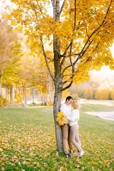 Hombre abraza a mujer con corona de hojas amarillas cerca de árbol en bosque otoñal — Foto de Stock