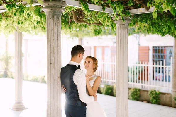 Жених и невеста стоят между колоннами под красивой зеленой лианой в старом городе, обнимаются и улыбаются — стоковое фото