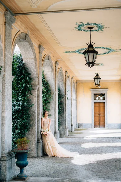 Beautiful bride in a dress with a bouquet of pink flowers is leaning against a pillar in the vaulted room. Lake Como, Italy — Stock Photo, Image