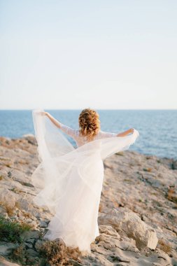 Gelin, kolları açık, kayalık bir deniz kıyısında duruyor. Arkasında ince bir etek kumaşı rüzgarda çırpınıyor. 