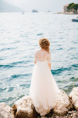 Zarif bir gelin kayalık bir deniz kıyısında duruyor, önünde eski kasaba ve tekneler, arka görüş. 