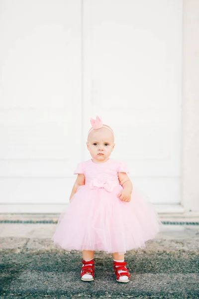 Une charmante petite fille en robe rose, baskets et avec une couronne sur la tête se tient à une porte blanche dans la vieille ville — Photo