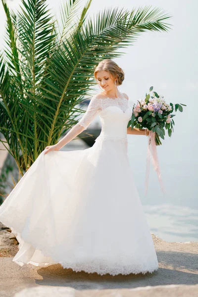 Una tenera sposa si trova vicino alla palma e tiene un bouquet da sposa e il bordo della gonna. — Foto Stock