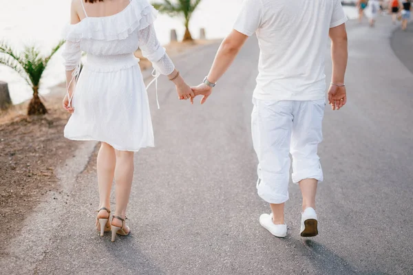 Een paar minnaars in witte kleren lopen langs de weg bij de zee, een vrouw houdt zachtjes een mannenhand vast — Stockfoto