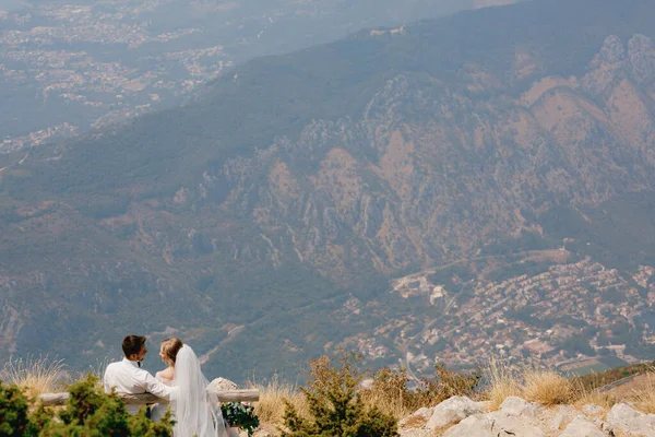 La novia y el novio se abrazan tiernamente y se sientan en un banco de madera en las montañas y se miran entre sí —  Fotos de Stock